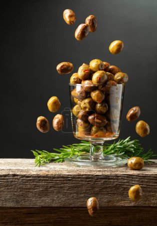 Foto de Aceitunas picantes en un tazón de vidrio. Cuenco con aceitunas conservadas y ramitas de romero sobre una mesa de madera. - Imagen libre de derechos