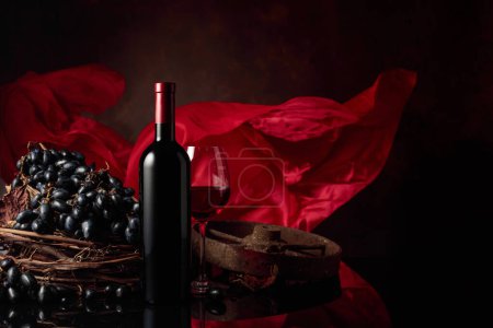 Photo for Glass and bottle of red wine with grapes on a black reflective background. Red satin curtain flutters in the wind. Copy space. - Royalty Free Image