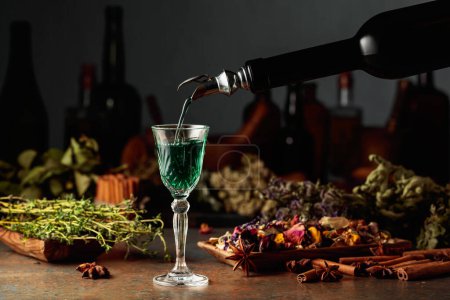 Photo for Green herbal liquor is poured from a vintage bottle into a glass. On a table dried herbs, flowers, spices, and old kitchen utensils. Concept of herbal medicine. - Royalty Free Image