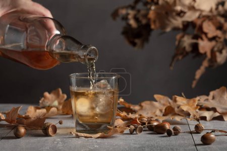 Téléchargez les photos : Le whisky est versé d'une bouteille dans un verre avec de la glace. Whisky en verre congelé avec glace sur une table en pierre grise avec des feuilles de chêne séchées. - en image libre de droit
