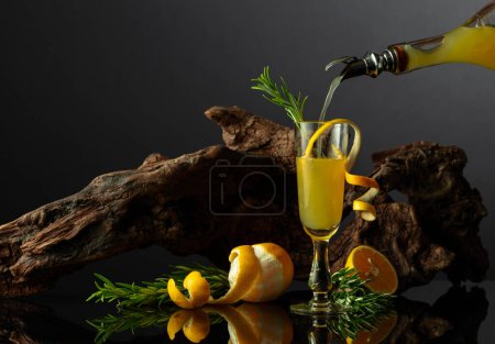 Téléchargez les photos : Liqueur italienne traditionnelle Limoncello sur fond noir réfléchissant. Verre de liqueur de citron avec brindille de romarin sur un fond de vieux chicots. L'alcool est versé dans un verre. - en image libre de droit