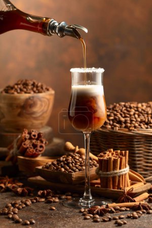Téléchargez les photos : La liqueur de café est versée d'une bouteille dans un verre avec de la crème. Des grains de café, des bâtonnets de cannelle et de l'anis sont éparpillés sur la table. - en image libre de droit