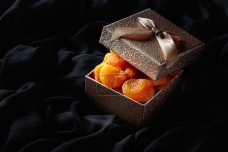 Téléchargez les photos : Abricots secs dans une boîte cadeau sur un chiffon noir. - en image libre de droit