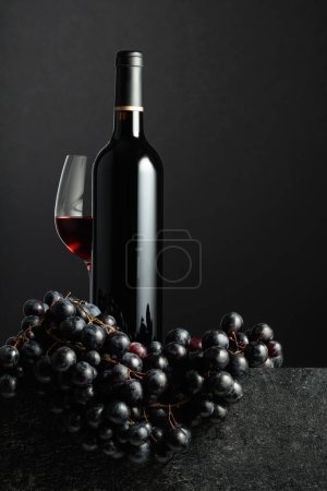 Téléchargez les photos : Juicy blue grapes and a bottle of red wine on a black background. - en image libre de droit