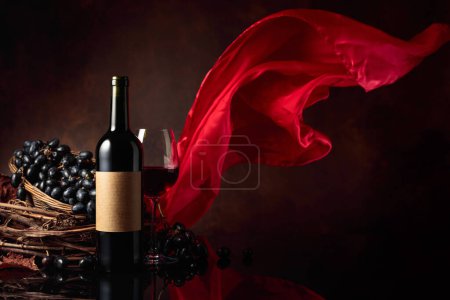 Téléchargez les photos : Verre et bouteille de vin rouge avec raisins sur fond noir réfléchissant. Sur une vieille étiquette vide. Rideau en satin rouge flotte dans le vent. Espace de copie. - en image libre de droit