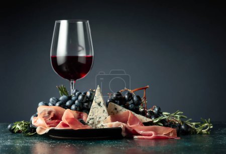 Téléchargez les photos : Vin rouge avec raisins, romarin, prosciutto et fromage bleu sur fond sombre. - en image libre de droit