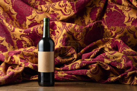 Photo for Bottle of red wine on a olld wooden tfble. In the background retro tapestry with dark red and golden floral ornament. - Royalty Free Image