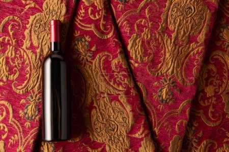 Foto de Botella de vino tinto sobre un tapiz retro con adorno floral rojo oscuro y dorado. Vista superior. - Imagen libre de derechos