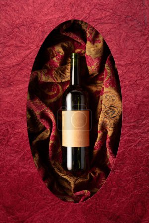 Téléchargez les photos : Bouteille de vin rouge sur une tapisserie rétro à l'ornement floral rouge foncé et doré. Sur une vieille étiquette vide. Vue du dessus. - en image libre de droit