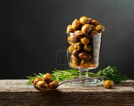 Foto de Aceitunas picantes en un tazón de vidrio. Cuenco con aceitunas conservadas y ramitas de romero sobre una mesa de madera. - Imagen libre de derechos