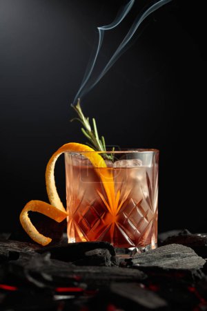 Photo for Old-fashioned cocktail with ice, orange peel, and rosemary. Whiskey with a burning twig of rosemary on a dark background. - Royalty Free Image