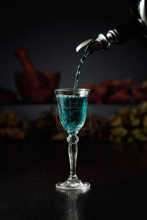 Téléchargez les photos : Le gin bleu est versé d'une bouteille vintage dans un verre. En arrière-plan herbes séchées et vieux ustensiles de cuisine. - en image libre de droit