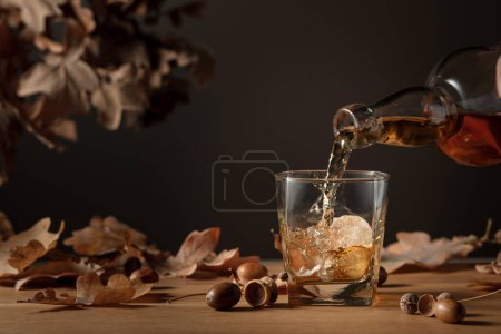 Foto de El whisky se vierte de una botella en un vaso con hielo. Whisky en vidrio congelado con hielo sobre una mesa de madera con hojas secas de roble. - Imagen libre de derechos