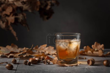Photo for Whiskey in frozen glass with ice on a grey stone table with dried-up oak leaves. - Royalty Free Image