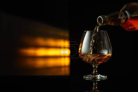 Photo for Pouring brandy from a bottle into a snifter on a black reflective background. Copy space. - Royalty Free Image