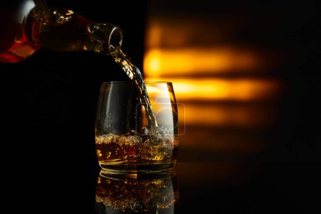 Photo for Pouring whiskey from a bottle into a glass on a black reflective background. Copy space. - Royalty Free Image