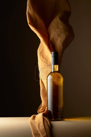 Foto de Botella de vino blanco y aleteos de tela beige sobre fondo oscuro. - Imagen libre de derechos