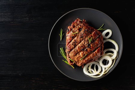 Photo for Grilled ribeye beef steak with rosemary and marinated onion on a black plate. Copy space. - Royalty Free Image