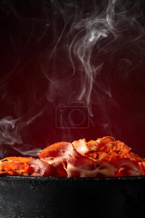 Photo for Fried steaming bacon slices in an old black pan on a dark red background. Copy space. - Royalty Free Image