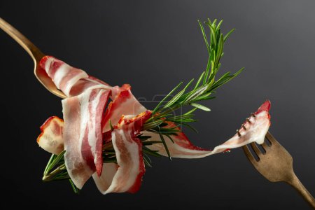 Photo for Dry-cured pork belly bacon with rosemary on a black background. Sliced bacon on a forks. - Royalty Free Image