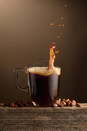 Téléchargez les photos : Espresso tasse en verre de café avec éclaboussures sur un fond brun. Tasse à café et grains de café torréfiés sur une vieille planche en bois. - en image libre de droit