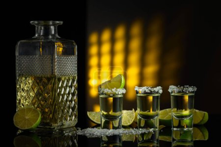 Téléchargez les photos : Tequila dorée avec sel de mer et tranches de citron vert sur fond réfléchissant noir. - en image libre de droit