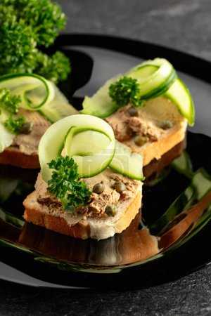 Photo for Open sandwiches with pate, fresh cucumber, capers, and parsley. Toasts with pate on a black plate. - Royalty Free Image