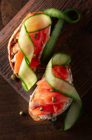 Photo for Sandwiches with smoked trout, cream cheese, fresh cucumber, and capers on an old cutting board. Top view. - Royalty Free Image
