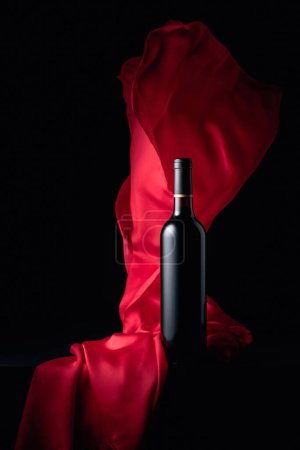 Foto de Botella de vino tinto y aleteos de tela roja sobre fondo negro. - Imagen libre de derechos