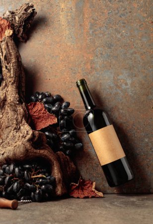 Photo for Bottle of red wine with blue grapes on a rusty background with an old snag and dried-up vine leaves. On a bottle old empty label. Concept of vinery. - Royalty Free Image