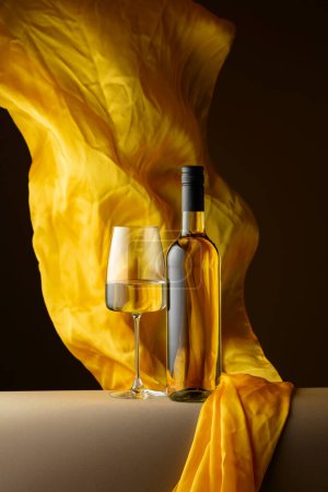 Foto de Botella y copa de vino blanco y aleteos de tela amarilla sobre fondo oscuro. - Imagen libre de derechos