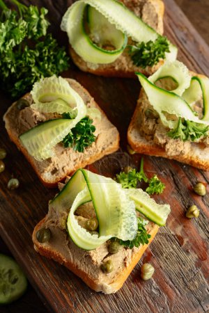 Photo for Toasts with pate on a old cutting board. Open sandwiches with pate, fresh cucumber, capers, and parsley. - Royalty Free Image