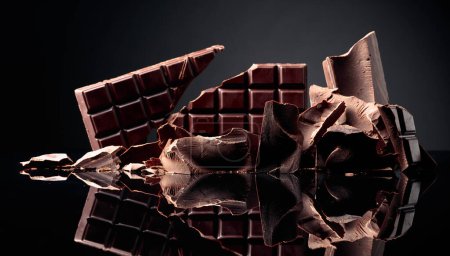 Photo for A broken chocolate bar and pieces of dark chocolate on a black reflective background. Copy space. - Royalty Free Image