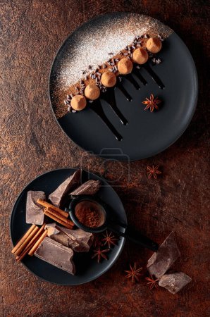 Photo for Chocolate truffles on a black plate with chocolate sauce. Sweets are sprinkled with cocoa powder and powdered sugar. Nearby are pieces of chocolate, cinnamon, and anise. - Royalty Free Image