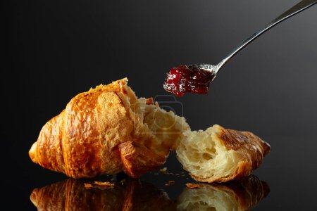 Foto de Croissant recién horneado con mermelada de frambuesa sobre un fondo negro reflectante. - Imagen libre de derechos