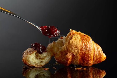 Foto de Croissant recién horneado con mermelada de frambuesa sobre un fondo negro reflectante. - Imagen libre de derechos