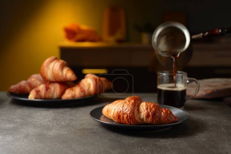 Téléchargez les photos : Croissants frais cuits au four et café noir sur une table de cuisine. Le café est versé dans une tasse. Concentration sélective. - en image libre de droit
