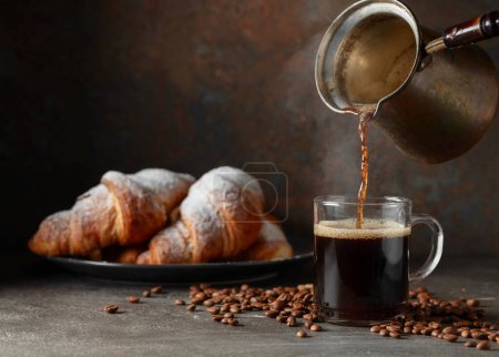 Foto de El café se vierte en una taza. Croissants y café negro en una mesa de la cocina. Concéntrate en un primer plano. - Imagen libre de derechos