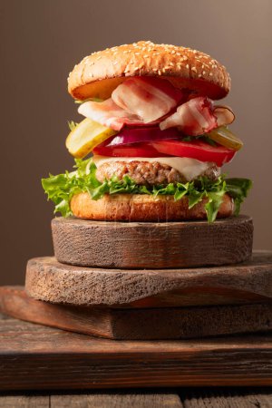 Téléchargez les photos : Hamburger frais savoureux sur une planche à découper en bois. Burger à la tomate, à l'oignon, au concombre en conserve, à la salade, au fromage, à l'escalope de bœuf et au bacon. - en image libre de droit