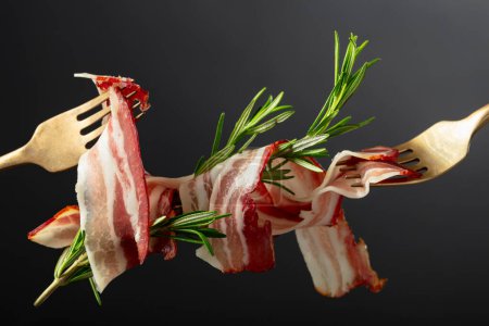 Photo for Dry-cured pork belly bacon with rosemary on a black background. Sliced bacon on a forks. - Royalty Free Image