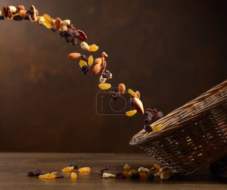 Foto de Frutas secas y frutos secos caen en la cesta. La mezcla de nueces secas y pasas sobre un fondo marrón. Copiar espacio. - Imagen libre de derechos