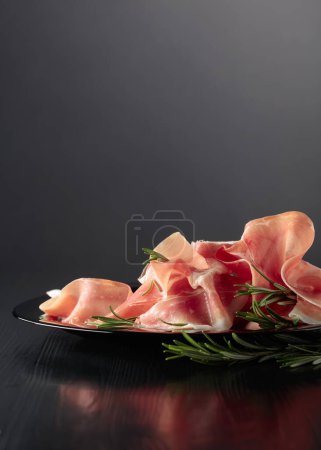 Photo for Italian prosciutto or Spanish jamon with rosemary on a black plate. Copy space. - Royalty Free Image