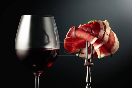 Téléchargez les photos : Verre de vin rouge et de prosciutto tranché avec romarin sur une fourchette, fond noir. - en image libre de droit