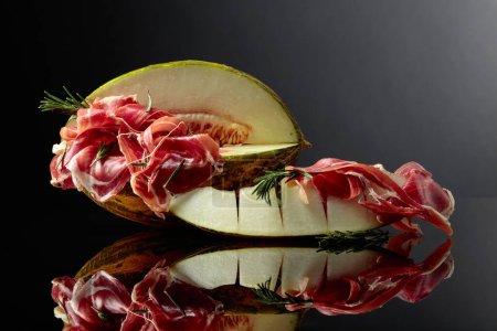 Photo for Prosciutto with melon and rosemary on a black reflective background. - Royalty Free Image