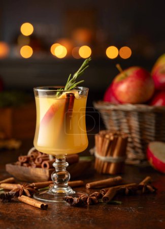 Photo for Mulled cider with slice apples, cinnamon, rosemary, and anise stars in glass cup on a brown rustic table. Holiday traditional hot drink. On a dark background kitchen utensils and burning candles. - Royalty Free Image