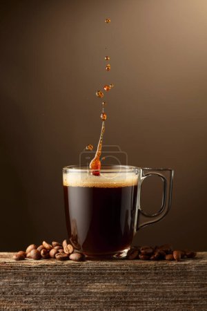 Téléchargez les photos : Espresso tasse en verre de café avec éclaboussures sur un fond brun. Tasse à café et grains de café torréfiés sur une vieille planche en bois. - en image libre de droit