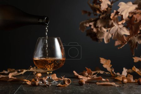 Foto de El brandy se vierte de una botella en un vaso. Francotirador de coñac sobre una mesa de piedra con hojas secas de roble. - Imagen libre de derechos