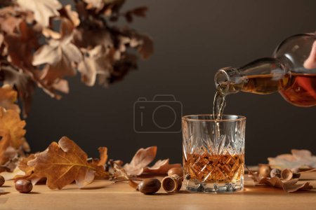 Téléchargez les photos : Le whisky est versé d'une bouteille dans un verre avec de la glace. Whisky en verre congelé avec glace sur une table en bois avec des feuilles de chêne séchées. - en image libre de droit