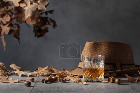 Photo for Whiskey in crystal glass on a grey stone table with dried oak leaves and a man's hat. - Royalty Free Image
