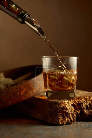 Foto de El whisky se vierte de una botella en un vaso congelado con hielo natural. Fondo oxidado áspero. - Imagen libre de derechos